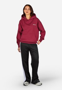 Bordová mikina s kapucňou a predným vreckom, s logom "Cozy Club", v kombinácii s čiernymi teplákmi so striebornými bočnými prúžkami a striebornými teniskami.