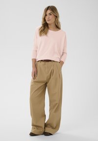 Pull rose pâle surdimensionné avec manches côtelées, associé à un pantalon kaki à plis taille haute. Ceinture marron et chaussures foncées complètent la tenue.
