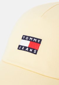 Sárga sapka bordázott textúrával, tűzött logócéggel, piros, fehér és tengerészkék színekben, amelyen a "TOMMY JEANS" felirat olvasható.