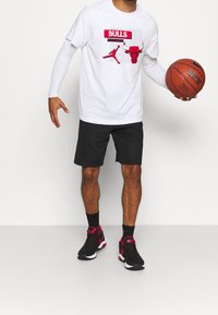 Camiseta blanca con gráfico rojo de Chicago Bulls y logo de Jordan, pantalones cortos negros y zapatillas de baloncesto negras/rojas. Sosteniendo un baloncesto.