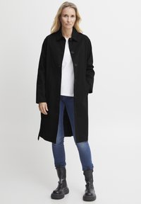 Melna, gara mēteli ar taisnu griezumu, pogu aizdari un sānu kabatām, apvienojot ar ziliem džinsiem un melniem platformas zābakiem.