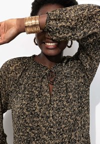 Blouse noire à motifs floraux avec un col froncé et un détail de lien. Manches longues avec des poignets élastiques. Modèle portant des bracelets en or et des boucles d'oreilles en créoles.