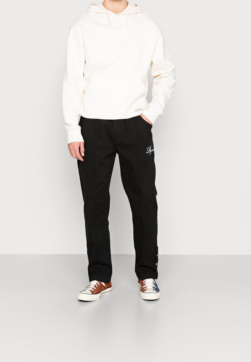 Personne portant un sweat à capuche blanc uni, un pantalon noir avec un petit logo blanc sur la cuisse gauche, et des baskets marron et bleues dépareillées.