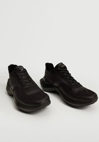 Chaussures de sport noires avec une tige en mesh, une surface texturée, un bout arrondi et une semelle rembourrée. Dotées de lacets et d'un détail de marque minimal.