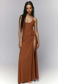Vestido maxi marrón con un corpiño ajustado, tirantes finos y un detalle de torsión con un acento circular en la cintura, que presenta una abertura alta en el muslo.