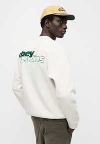 Λευκό φούτερ με πράσινο λογότυπο "obey" και το κείμενο "Studios" στην πλάτη, σε συνδυασμό με ένα κίτρινο καπέλο με μαύρη κέντηση.