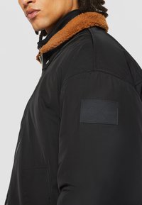 Doudoune noire avec col en fourrure beige, dotée d'un patch sur le bras gauche en relief avec "Calvin Klein Jeans". Texture lisse et forme structurée.