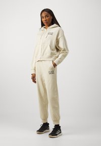 Bežna zip-up kapuca s prednjimi žepi in logotipom, kombinirana z usklajenimi tapered jogging hlačami, ki imajo vezen logotip. Črne športne superge.