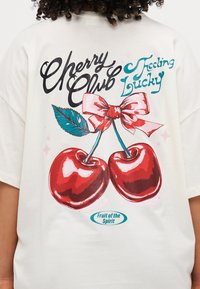 Valge oversized T-särk, millel on graafiline kujundus, kus on kaks punast kirssi, roosa pael ja lehed. Tekst ütleb "Cherry Club Feeling Lucky."