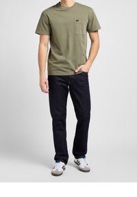 Camiseta de manga corta verde oliva con un bolsillo en el pecho, combinada con unos jeans azul oscuro y zapatillas blancas con rayas negras y acentos grises.