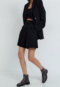 Blazer nero sartoriale con un singolo bottone blu, abbinato a pantaloncini neri a vita alta e stivaletti grigio lucido con cerniere.