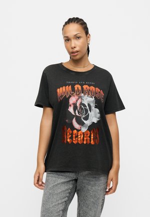 Kobieta z zaplecionymi włosami ma na sobie czarny t-shirt z "Wild Rose Records" i grafiką róży, do tego jasnoszare dżinsy, stoi neutralnie.