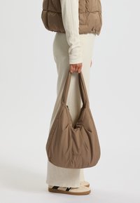Bolso de hombro marrón, relleno y de nylon, con una textura suave y correas anchas. Incluye un sutil logotipo en la parte frontal.