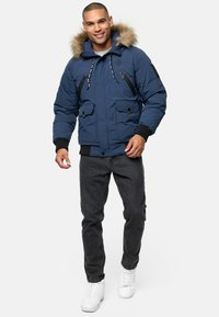 Marineblå polstret jakke med pelsforet hætte, to frontlommer med klap, lynlåslomme på brystet og ribbede manchetter. Båret over mørke jeans og hvide sneakers.