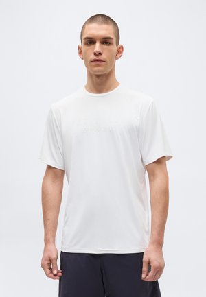 Jeune homme aux cheveux courts portant un t-shirt blanc uni à manches courtes avec un logo discret en relief, debout devant un fond clair.