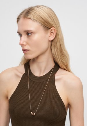 Blonde Frau mit blauen Augen, die ein ärmelloses, dunkelbraunes Rippenoberteil und eine zarte Halskette mit drei goldenen Perlen trägt und nach links schaut.