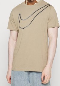 Khaki bavlněné tričko s černým logem Nike na přední straně, kulatým výstřihem a krátkými rukávy. Tkanina má jemnou strukturu.