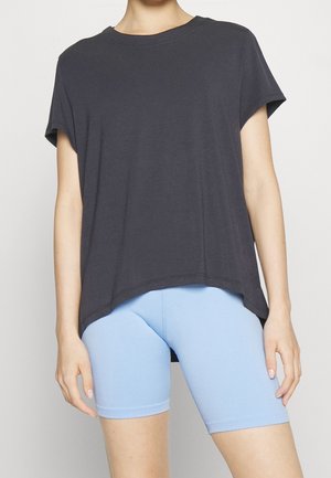 Osoba ubrana w luźną ciemnoszarą koszulkę z krótkim rękawem i dopasowane jasnoniebieskie spodenki rowerowe na jednolitym tle.