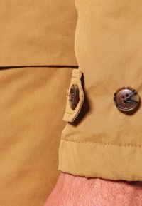 Ljusbrun bomullsduk med en slät textur har en knappad manschettdetalj som visar en rund, mönstrad knapp.