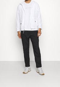Chaqueta blanca ligera con capucha, con bolsillos frontales con cremallera, lleva puesta una camiseta blanca con texto. Pantalones negros y zapatillas gris completan el conjunto.