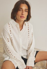 Camicia bianca in cotone con dettagli di ricamo a taglio intricati lungo le maniche e i lati, con una vestibilità rilassata e abbottonatura frontale.