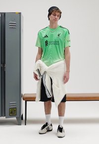 Zelený futbalový dres s čiernymi akcentmi, čierne šortky, biely uterák omotaný okolo pásu, čierne Adidas topánky, stojace pri sivom šatníku.