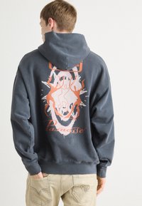 Sudadera gris con capucha, con un diseño gráfico en naranja y blanco en la parte posterior, que presenta formas abstractas y la palabra "Paradise".