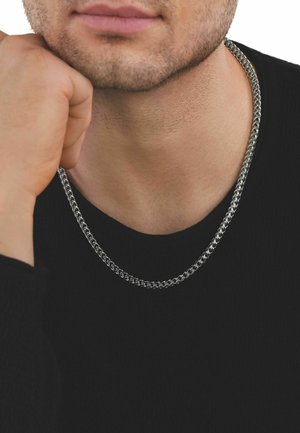 Bearded man wearing a silver chain necklace and black shirt, resting his chin on his hand.