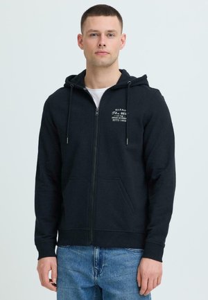 Jauns vīrietis, tērpies melnā rāvējslēdzēja džemperī ar baltu uzrakstu krūtīs un zilās džinsās, stāv pret vienkāršu gaišu fonu.