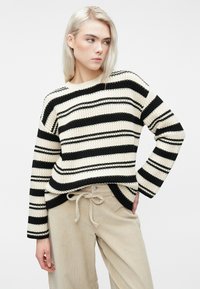 Noor blonde juustega noor inimene kannab kreemjas ja mustris triibulist kampsunit ning beeže pükse, seistes ühe käega puusal valge tausta Against.