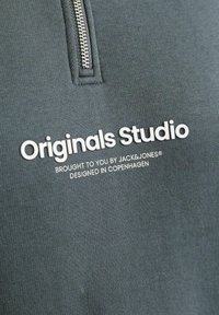 Materialul unui hanorac gri închis, cu o suprafață texturată, având un guler cu fermoar pe un sfert și un branding îndrăzneț cu text alb pe față.
