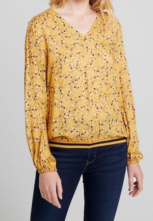 Femme portant un chemisier à manches longues jaune à fleurs avec des poignets élastiques et un jean bleu foncé sur un fond clair uni.