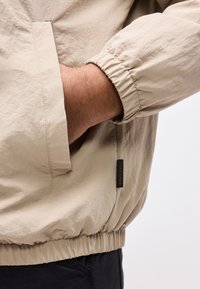 Χέρι ατόμου μέσα στην τσέπη μπεζ μπουφάν με ελαστικό μανίκι και εμφανές ετικέτα "Jack & Jones", φοράει μαύρο παντελόνι.