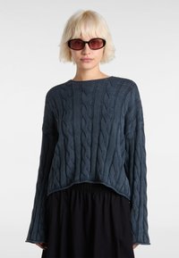 Navy blue cable-knit sweater with long sleeves and a round neckline. Soft texture and slightly cropped design, paired with a black skirt.