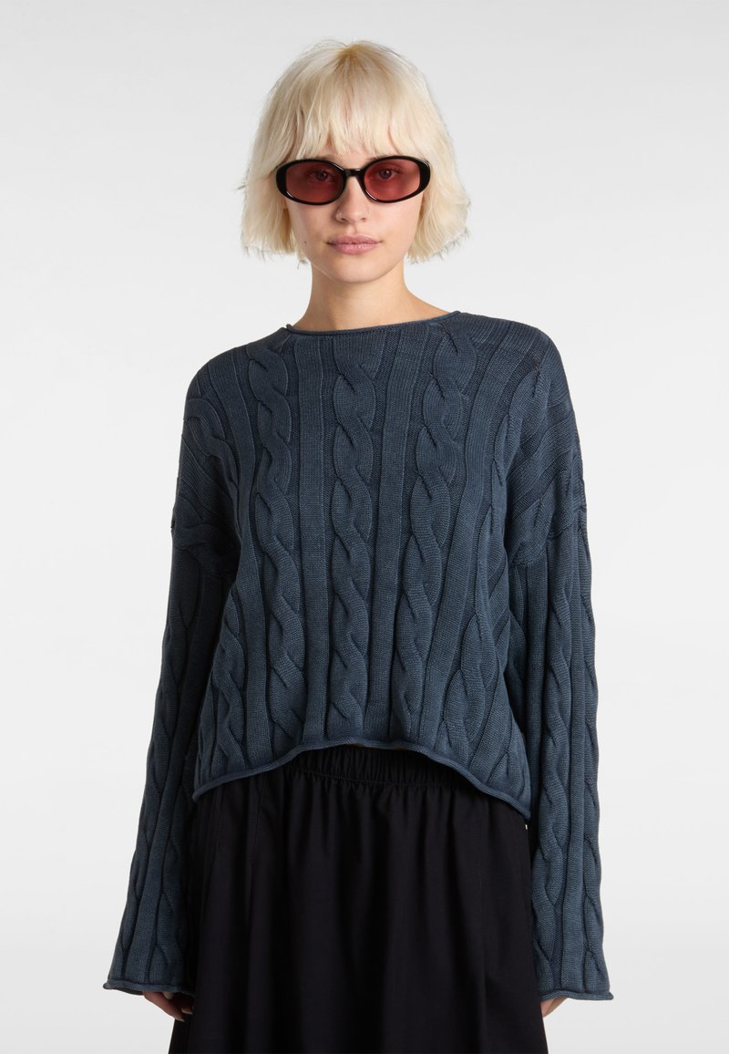 Navy blue cable-knit sweater with long sleeves and a round neckline. Soft texture and slightly cropped design, paired with a black skirt.