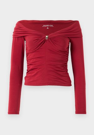Röd långärmad topp med off-the-shoulder-design, rynkade detaljer fram och en gulddetalj i mitten. Slät tygtextur.