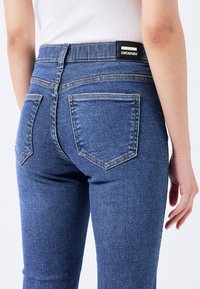 Vaqueros de mezclilla azul con una cintura alta, que cuentan con cinco bolsillos, costuras visibles y una etiqueta de marca negra en la parte posterior.