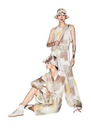 Deux femmes en tenues aux motifs abstraits atténués et chapeaux bob assortis ; l'une debout avec un sac bandoulière, l'autre assise avec des baskets et des chaussettes blanches.