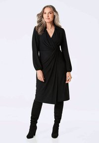 Schwarzes Wickelkleid mit langen Ärmeln, V-Ausschnitt und gerafftem Seitenelement. Kombiniert mit kniehohen schwarzen Stiefeln. Glatte Stofftextur.