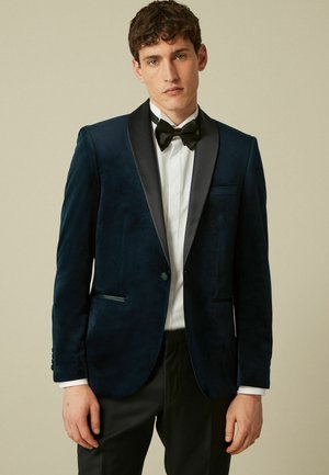 Marineblauer Samt-Tuxedo-Jacke mit satinierten Revers, Ein-Knopf-Verschluss und zwei frontalen Taschen. Getragen über einem weißen Hemd und einer Fliege.