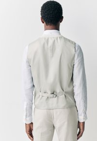 Gilet gris clair en tissu lisse, avec un dos ceinturé, porté par-dessus une chemise blanche à col, à manches longues et avec des accents de boutons.