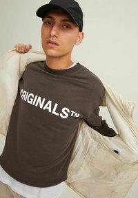 Brown long-sleeve shirt with "ORIGINALS™" text in white, worn under a light-colored jacket. The model holds the jacket open.
