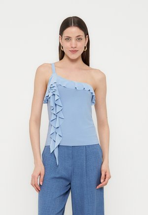 Γυναίκα με μακριά καστανά μαλλιά, ντυμένη με ένα γαλάζιο τοπ με έναν ώμο και βολάν και μπλε ψηλόμεσο παντελόνι σε απλό φόντο.