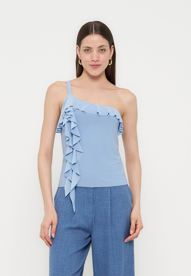 Γυναίκα με μακριά καστανά μαλλιά, ντυμένη με ένα γαλάζιο τοπ με έναν ώμο και βολάν και μπλε ψηλόμεσο παντελόνι σε απλό φόντο.