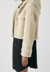 Osoba ubrana w skróconą beżową kurtkę sherpa z napami, nosząc czarną sukienkę na guziki, pokazana z boku na jednolitym tle.