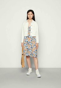 Jovem mulher vestindo uma jaqueta branca, um vestido florido até aos joelhos, tênis brancos, segurando uma bolsa de vime, em pé contra um fundo claro e liso.