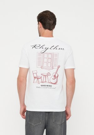 Άνδρας που φοράει λευκό T-shirt με σκίτσο παραθύρου, καρέκλας, τραπεζιού με κούπα, κιθάρας και κείμενο για τον ρυθμό και τη καλή μουσική στην πλάτη.