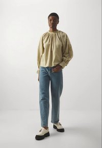 Blusa beige de manga larga con escote fruncido y mangas texturizadas, combinada con unos vaqueros rectos de un azul claro y unos zapatos gruesos en color marfil.