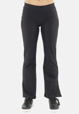 Personne portant un pantalon flare noir et ajusté ainsi que des baskets noires à semelles blanches, debout devant un fond clair uni.