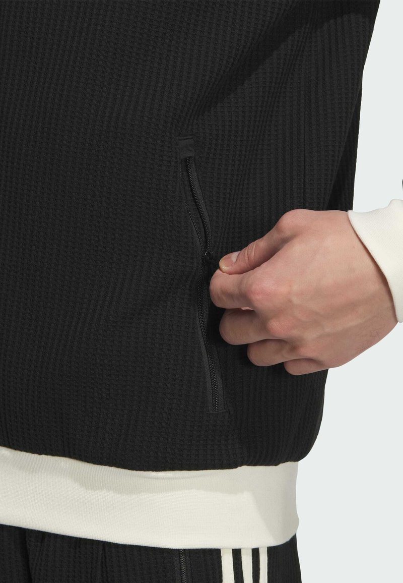 Chaqueta deportiva negra con textura que presenta un bolsillo con cremallera en el lateral y un dobladillo blanco contrastante. La tela de punto waffle añade profundidad.